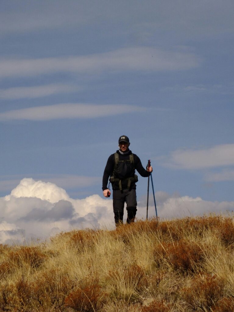 Man hiking with trekking poles over grassy terrain under a blue sky.